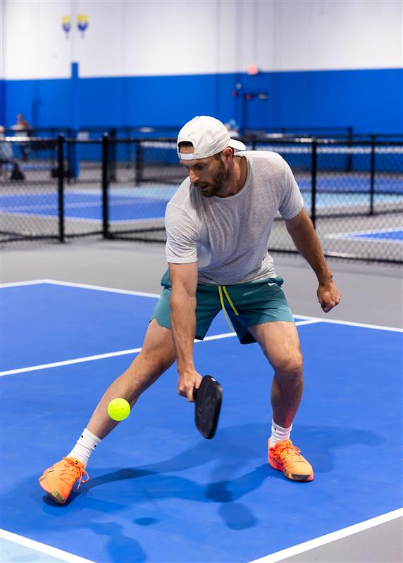 Two people playing pickleball on an indoor court, both focused on the game. The background shows a blue wall and a net. Text in image: Pickleball Kingdom. The mood is energetic and welcoming.