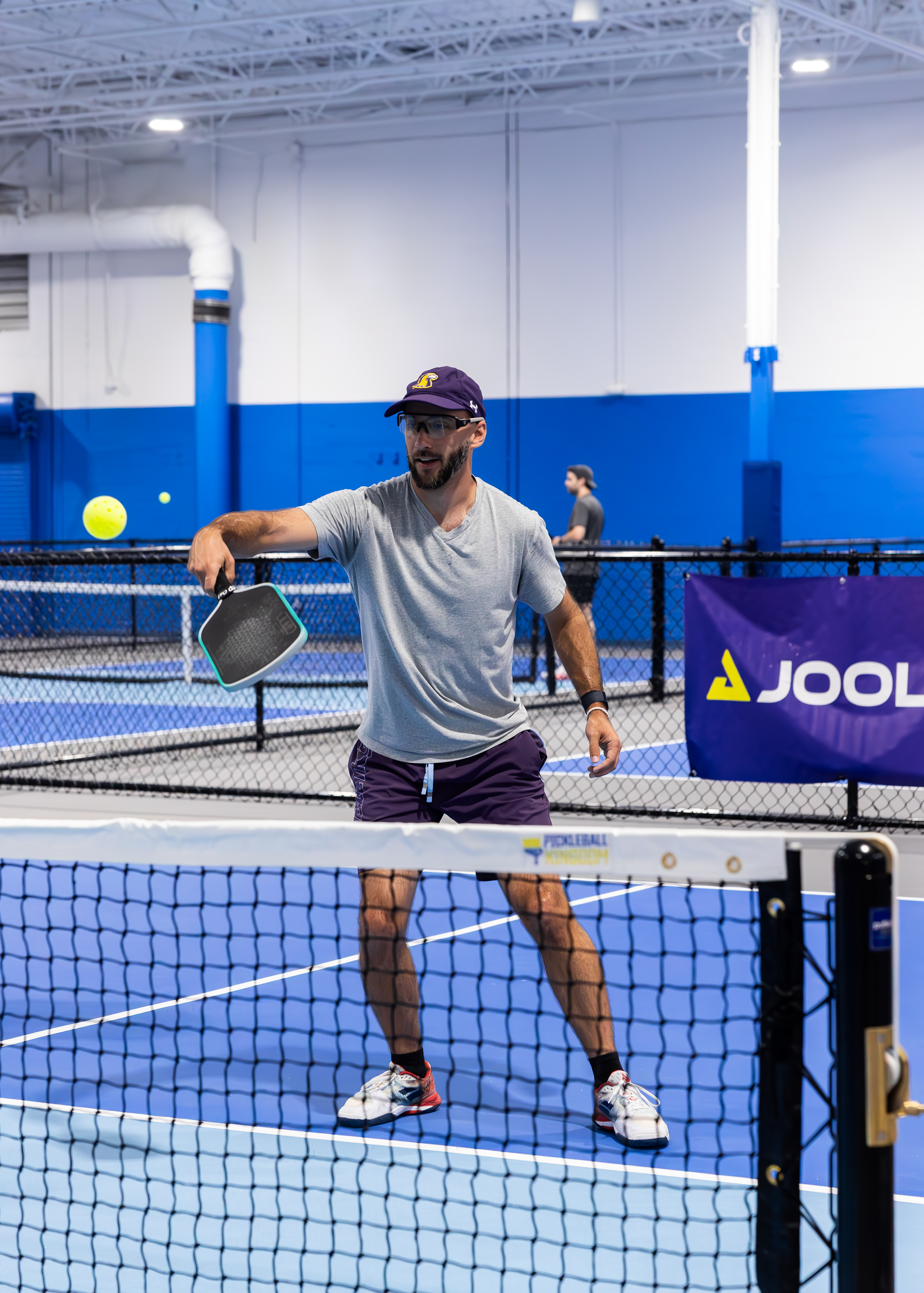 Two people playing pickleball on an indoor court, both focused on the game. The background shows a blue wall and a net. Text in image: Pickleball Kingdom. The mood is energetic and welcoming.