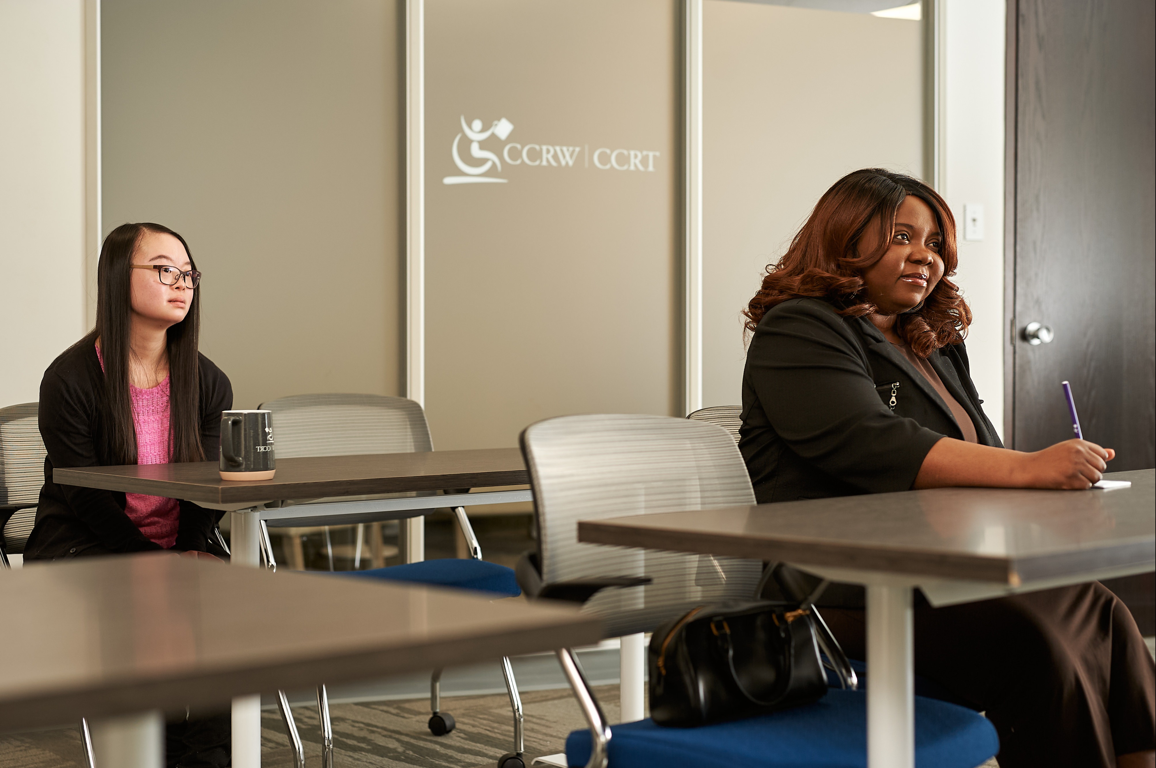 Two people in a facilitation room with the CCRW logo on a glass wall behind them. Both people are looking forward and one person is taking notes.