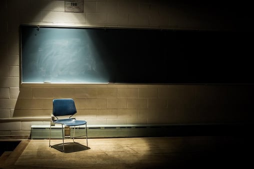 A single plastic chair sits in an empty classroom, lit up by a spotlight with an empty blackboard behind it.