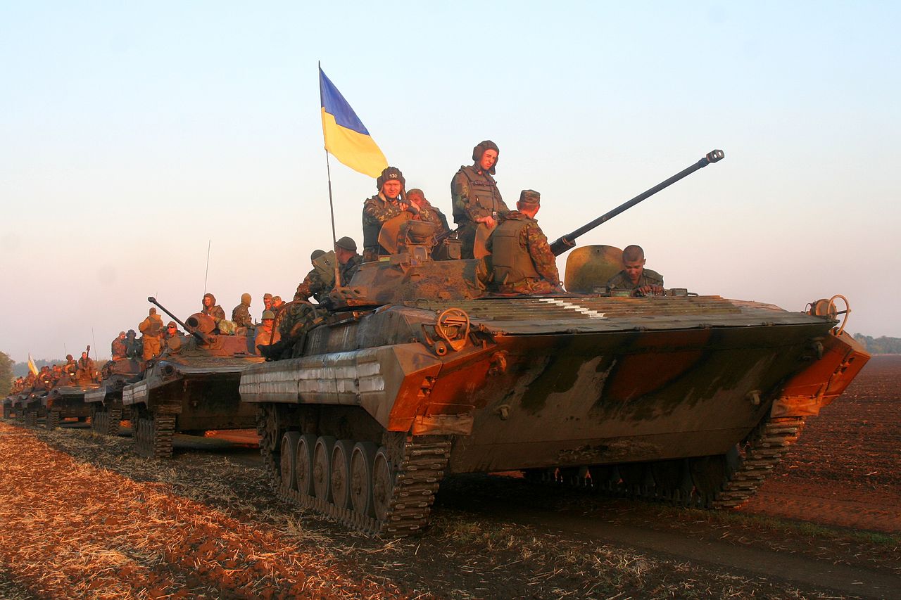A row of armored vehicles with men in combat gear sitting on top stretches into the distance as they drive over a field under a clear sky The front vehicle has a Ukrainian flag