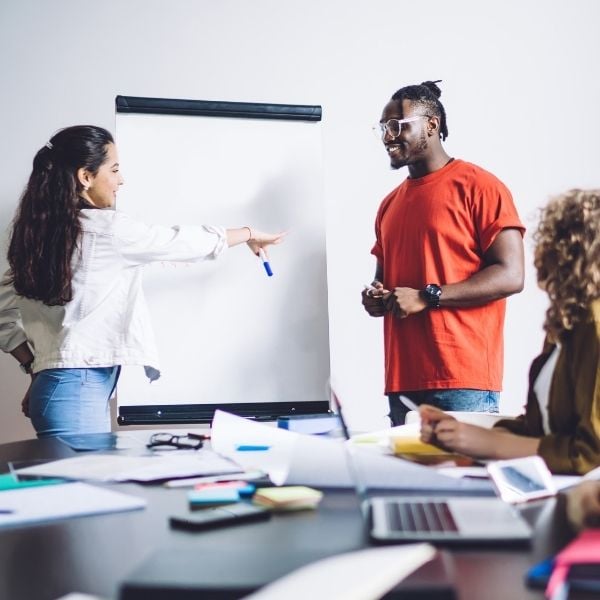 Two people engage in a simulated discussion while two other figures sit watching from a desk containing a laptop, notebooks and papers. 