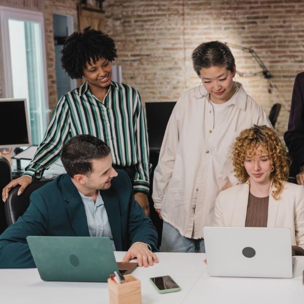 Two people site around a laptop, talking about it's content while 2 more of their colleagues stand behind them in an open-concept office space with a brick wall, and office furniture in the background.