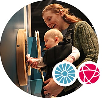 A woman holds her baby in a carrier on her front. She holds the baby's hand, helping them touch a textured display on the wall.
