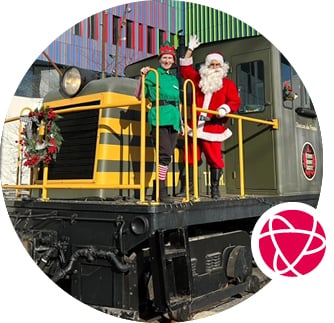 Santa and an elf wave while standing on the front of a locomotive decorated for the season.