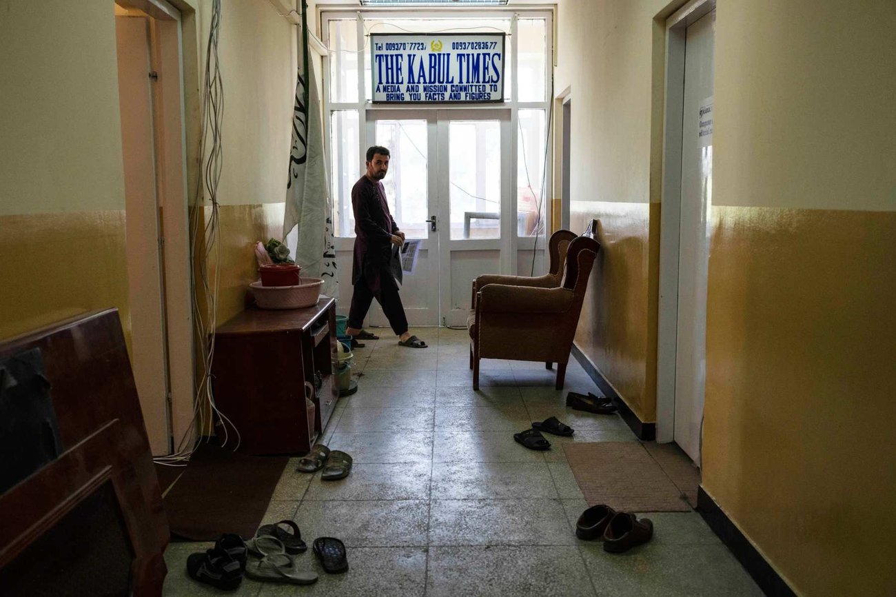 A hallway in a building with yellow walls and tiled floor. A man walks past a sign that read The Kabul Times