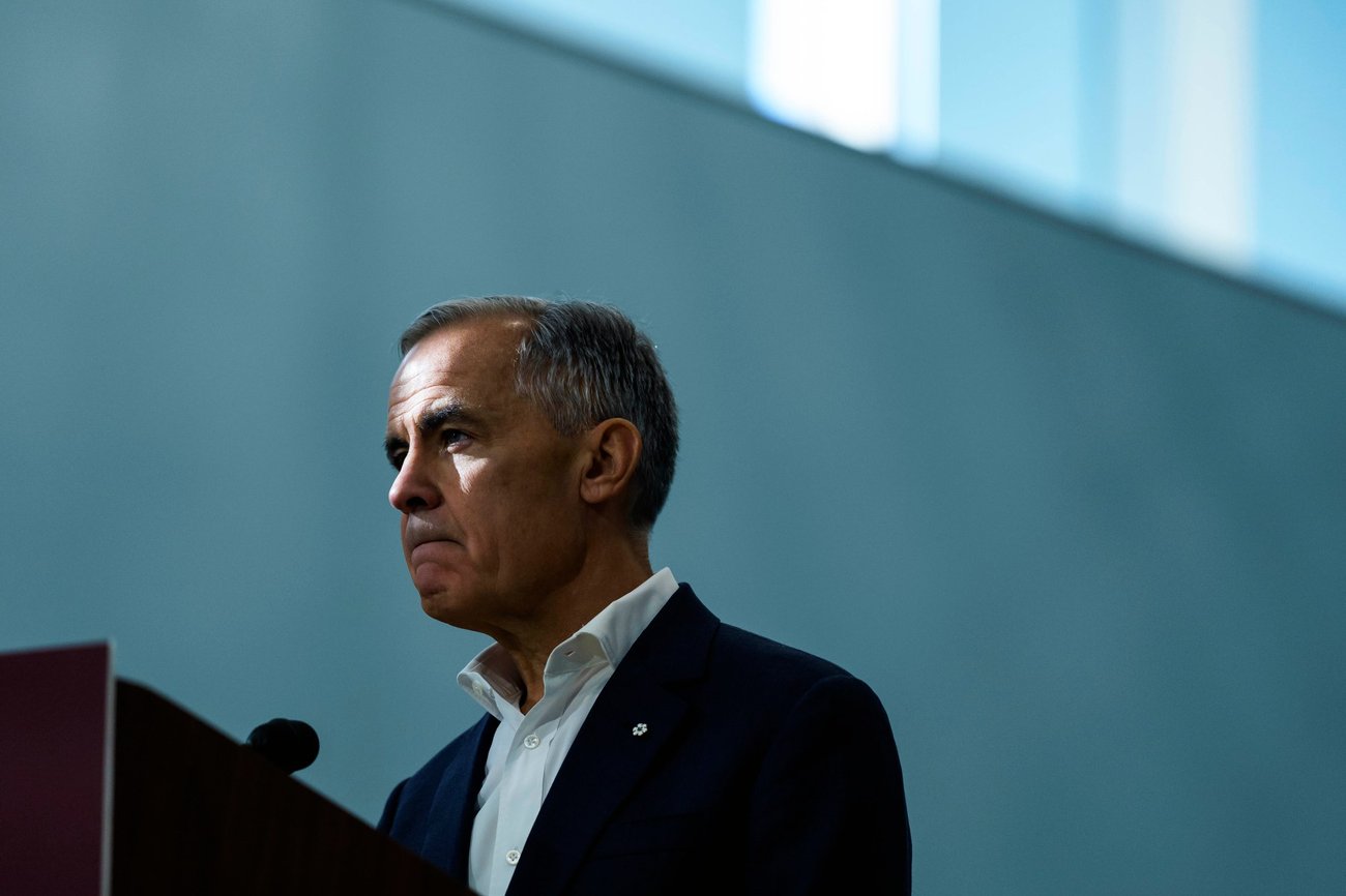 A photo of Prime Minister Mark Carney standing in front of a podium with a blue-grey backdrop behind him.