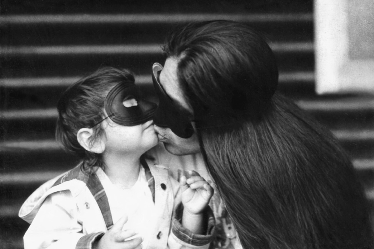Black and white photo of a person with long hair kissing a young boy. Both are wearing black costume masks over their eyes.