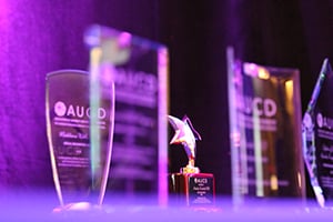 This image shows a collection of awards displayed on a surface. The focus is mainly on a star-shaped trophy in the center, which appears to be made of a reflective material with a gold and silver finish. Surrounding the star-shaped trophy are several transparent glass or acrylic plaques, each bearing text and logos, although the text is not fully legible due to the lighting and focus. The lighting has a purple hue, creating a vibrant and celebratory atmosphere. The awards appear to be from an organization abbreviated as "AUCD."
