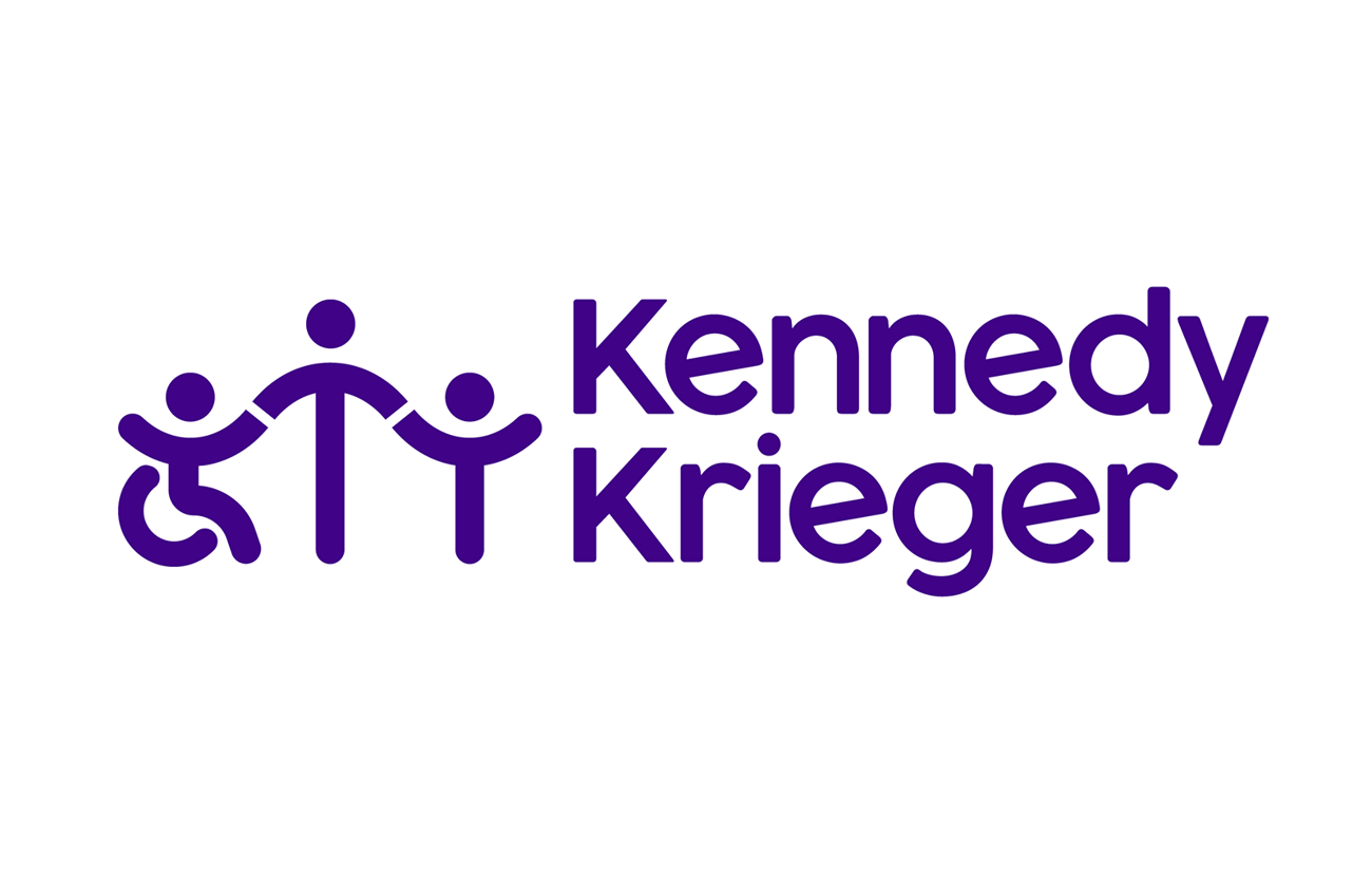 Kennedy Krieger Institute logo featuring three stylized human figures holding hands. The left figure is in a wheelchair, the center figure is taller, and the right figure is standing. All figures and the text 'Kennedy Krieger' are in purple.