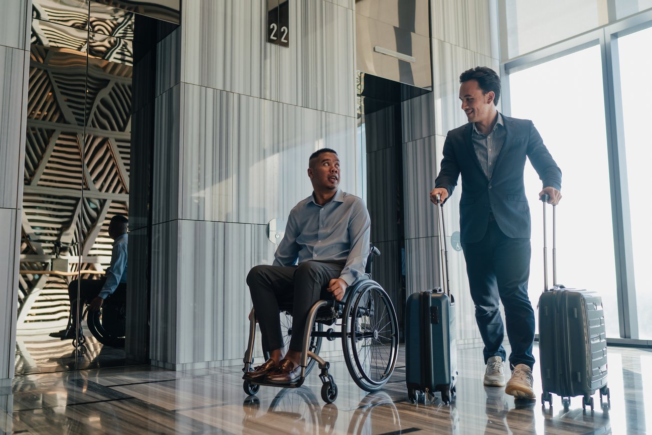 two travelling businessmen exit an elevator with luggage, one is in a wheelchair