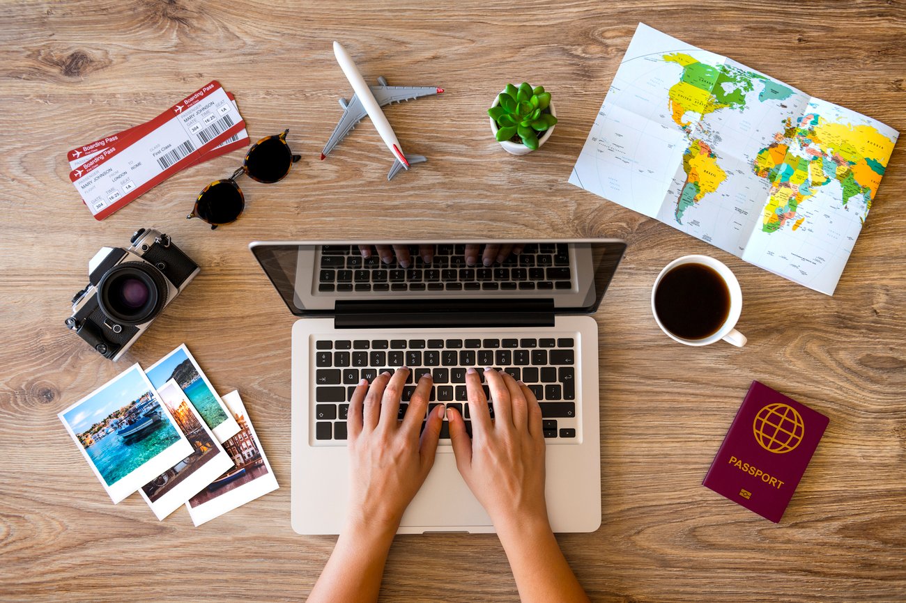  birds eye view of someone typing on their keyboard. There are several travel items around the table (map, sunglasses, tickets, passport, coffee, camera)