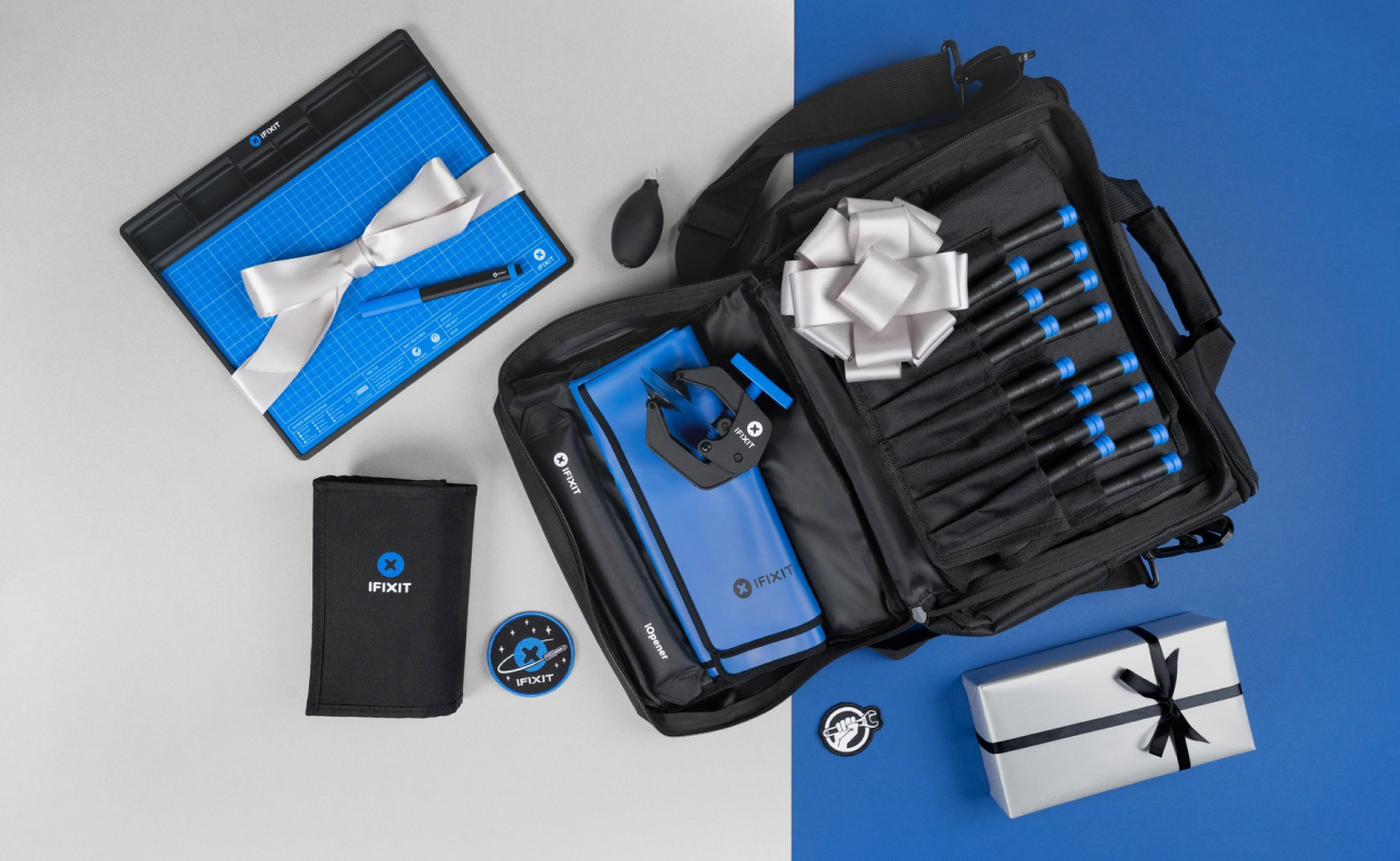 A flat lay on a split blue and gray background featuring the Repair Business Toolkit opened to display organized storage pockets, precision drivers, and repair accessories. A wrapped gift box sits beside the kit along with the FixMat and Pro Tech Toolkit, set against a dual-tone grey and blue background with a clean, festive layout.