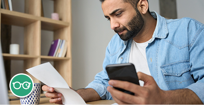 Man sitting at a desk, holding a smartphone in one hand and reading a sheet of paper, with shelves and a coffee cup in the background. In the bottom-left corner there is a small illustration of eyeglasses.