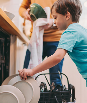 A child loading a white plate into a dishwasher. In the background, an adult is drying a green bowl with a dish towel.