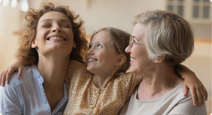 This photo is a picture of a mom, daughter, and grandma all smiling. The daughter is in the middle with her hands wrapped around both the mom and grandma