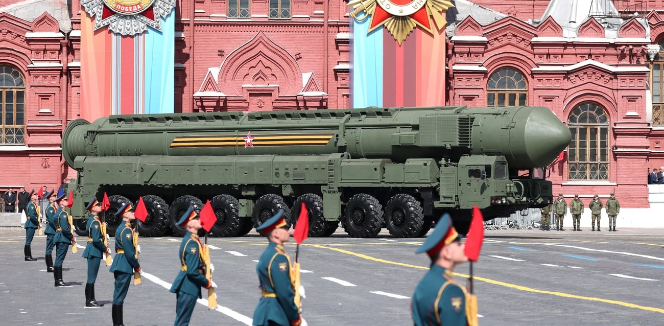Russian soldiers stand at attention as a large green missile on a multi-wheeled based is paraded through a street, with ornate brick buildings in the background.