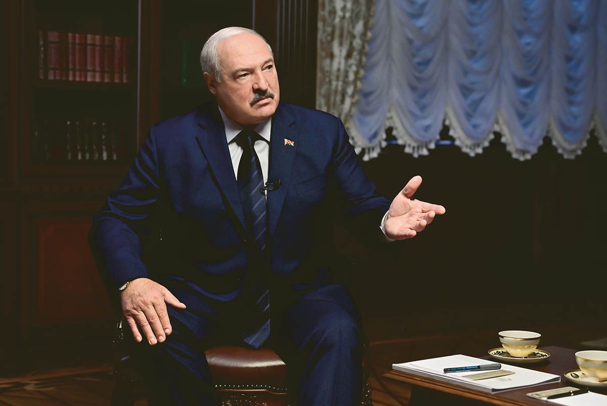 A man in sitting in a chair wearing a blue suit gestures out toward the right side of the photo