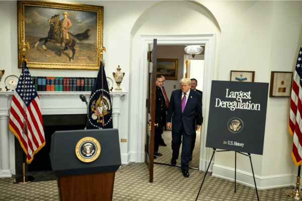 US President Donald Trump and others walk into a room setup for a press conference with a podium under a portrait of past US President Theodore Roosevelt riding a horse A sign says largest deregulation