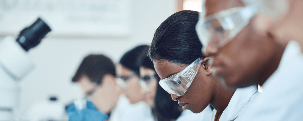 The faces of five students of diverse ethnicities looking down into microscopes while wearing protective glasses and lab coats. The faces of five students of diverse ethnicities looking down into microscopes while wearing protective glasses and lab coats.