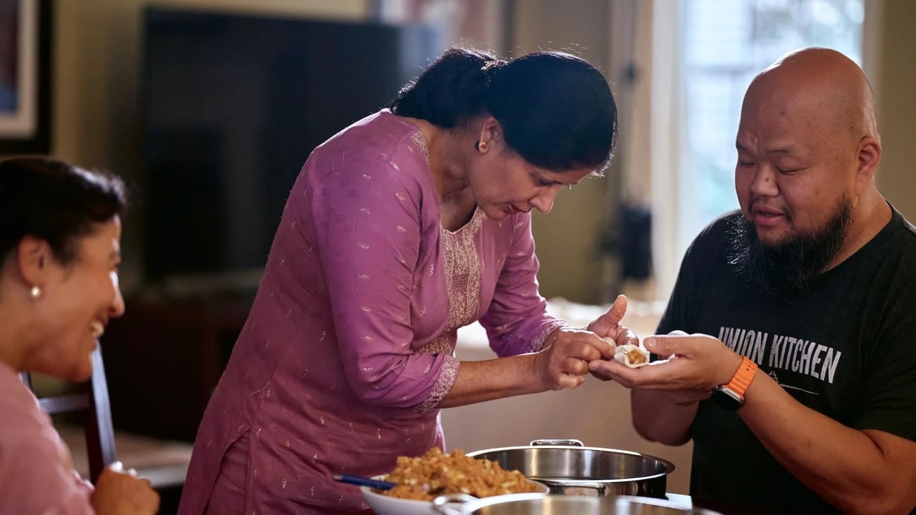 A woman helps a man fold a dumpling at a table while another woman smiles at them