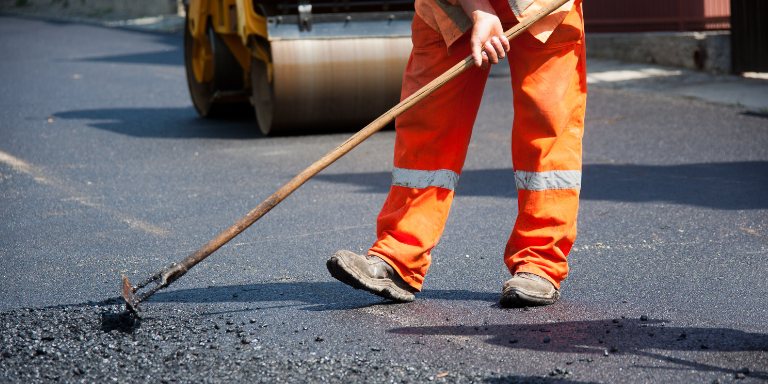 Photo of a person in orange safety clothing paving a road.