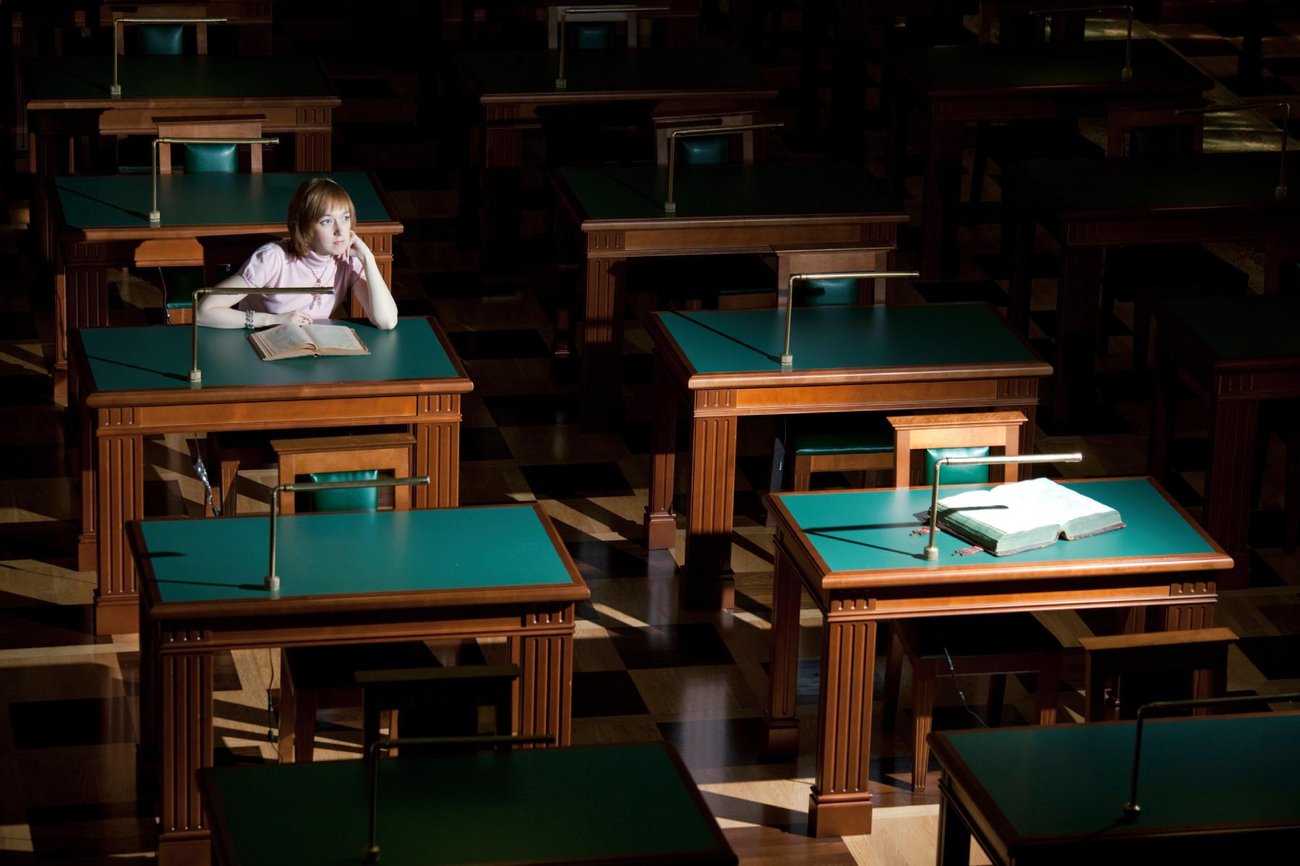 A woman sits at a green desk, with a book open in front of her, amidst two rows of similar desks, with light falling on only one of them while the others are in the dark