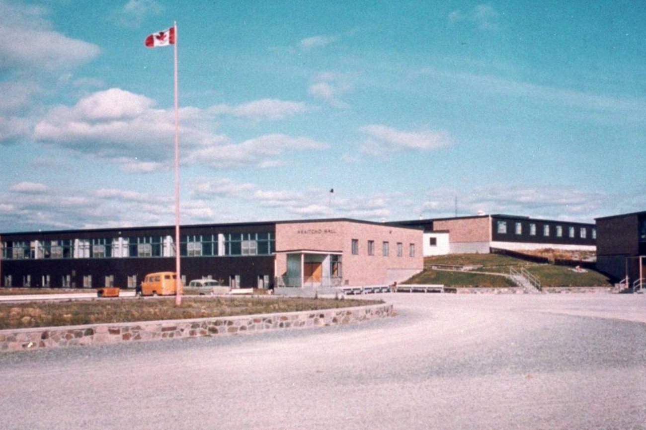 An old colour photograph showing a blue sky over several low school-like buildings. A Canadian flag is on the flag post