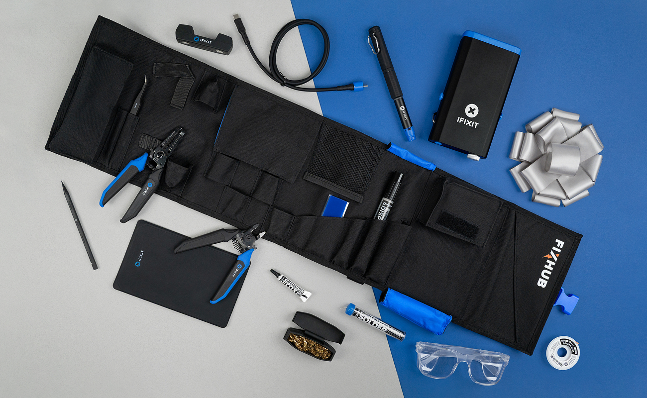 In-use lifestyle image showing the FixHub Soldering Toolkit roll fully opened on a grey-and-blue background, surrounded by iFixit pliers, precision tools, soldering accessories, safety glasses, and a wrapped silver bow.