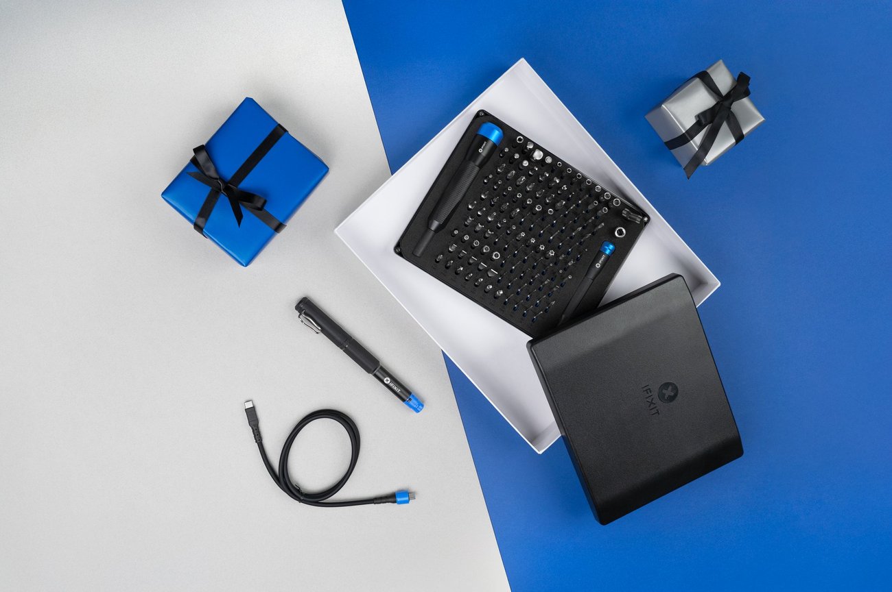 Promotional image showing the iFixit Manta Driver Kit opened inside a white gift box next to a FixHub Smart Soldering Iron on a blue-and-grey background.