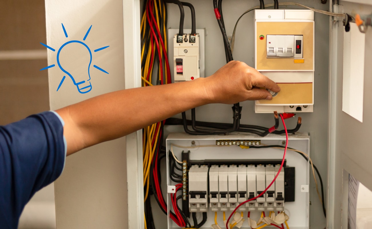 A close-up of a person’s hand adjusting a control inside an open electrical panel filled with wires and circuit breakers, with a blue lightbulb icon overlaid on the left side.