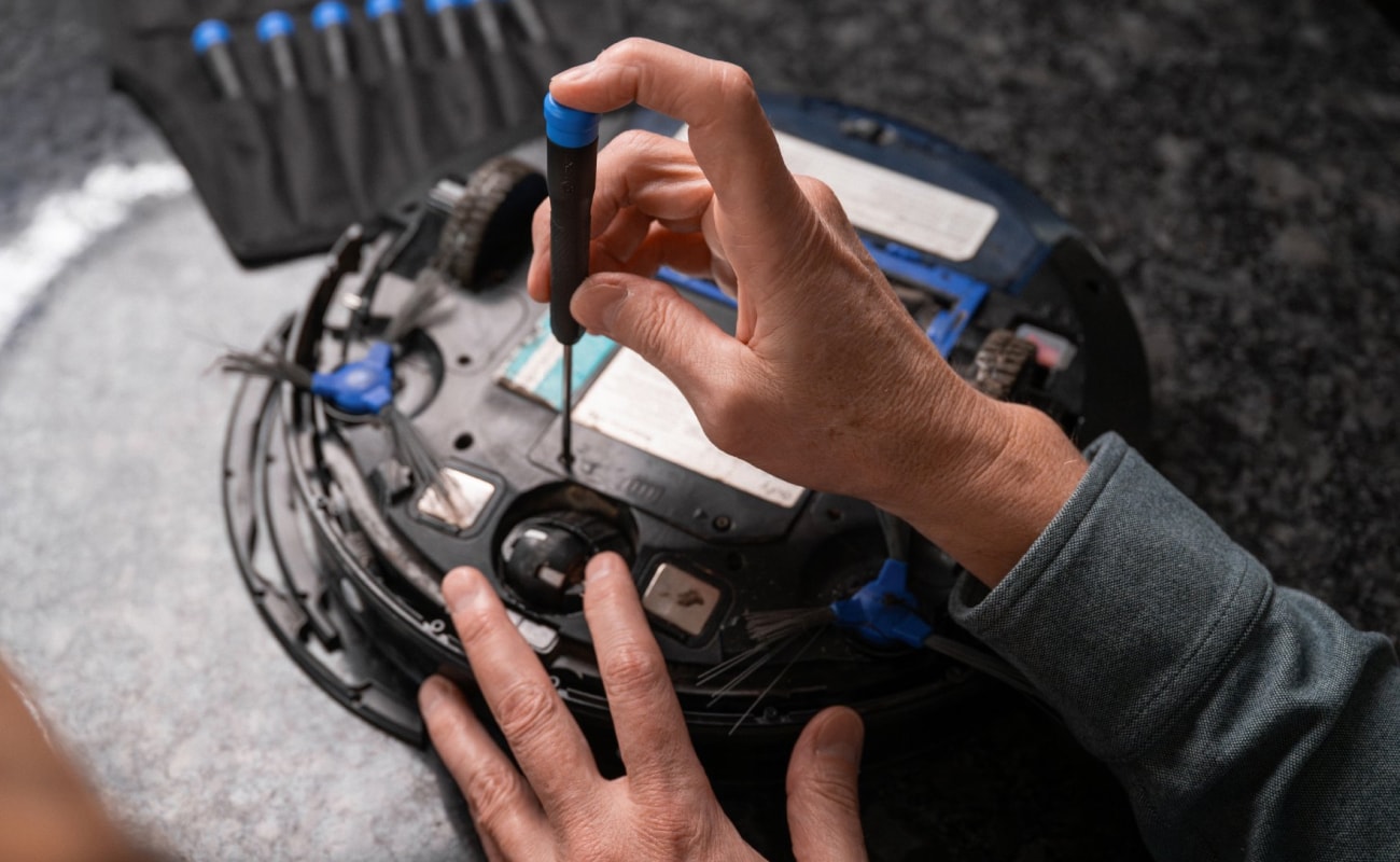 A close-up of hands using a precision screwdriver to open the underside of a robot vacuum, with tools and bits visible in the blurred background. The composition centers on the repair action, highlighting the device and the screwdriver in use.