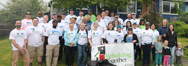  Aproximadamente 30 voluntarios con camisetas a juego de “Rebuilding Together South Sound” (Reconstruyendo South Sound juntos) se reúnen en el césped frente a una casa azul, sonríen y sostienen una pancarta. Se ven herramientas y equipos en primer plano.