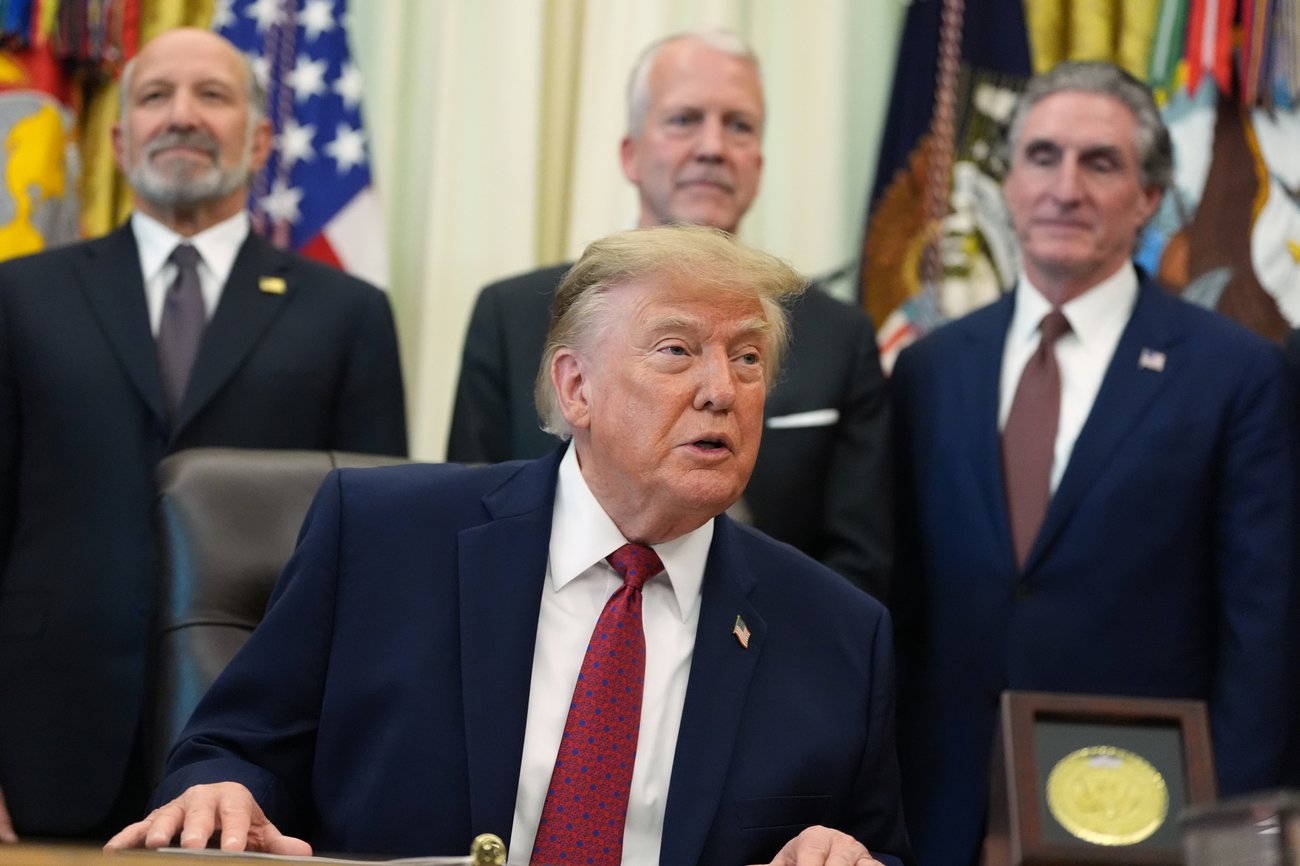 A suited man seated before flags with three suited men standing behind him