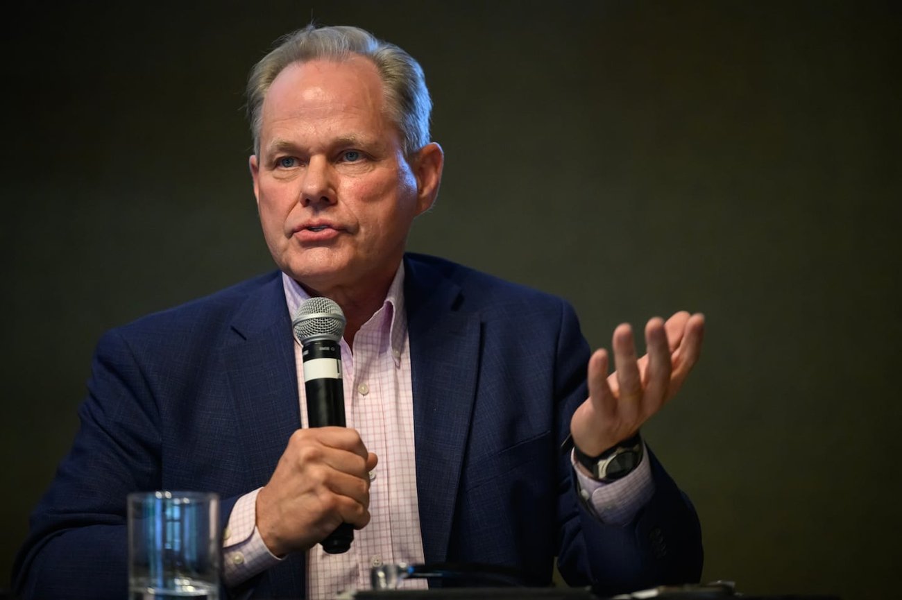 A man in a blazer speaks into a microphone while gesturing with one hand.