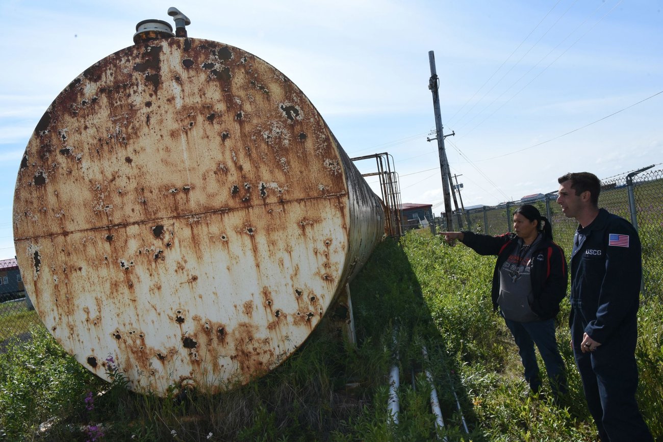 Two people stand beside a large rusted cylindrical tank outdoors