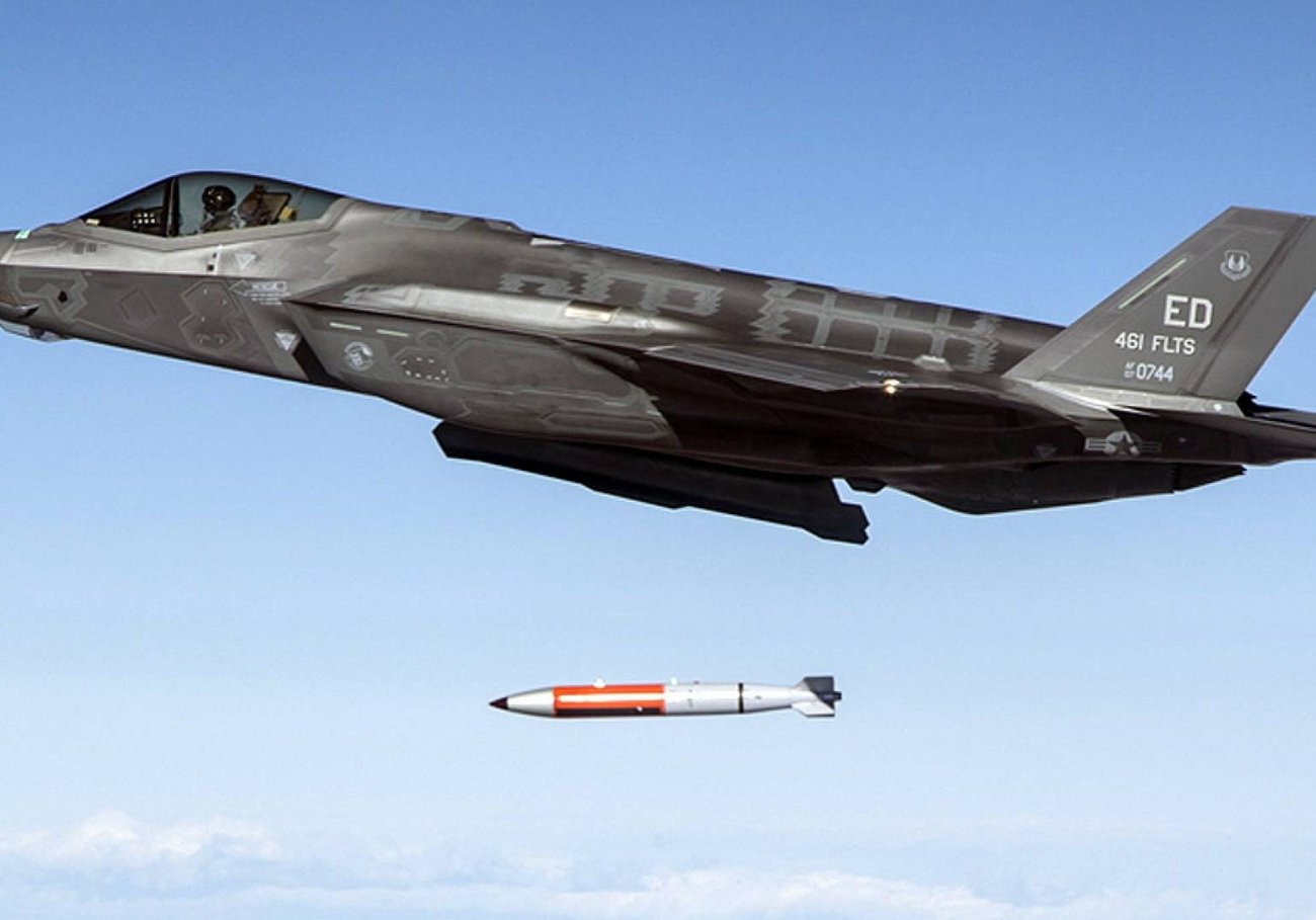 A black fighter jet drops an orange and grey bomb with a blue sky in the background.