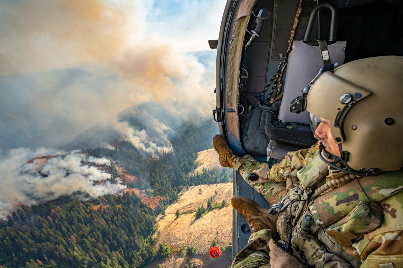 A man in military gear looks down from an aircraft over a forest on fire smoke rising into the air