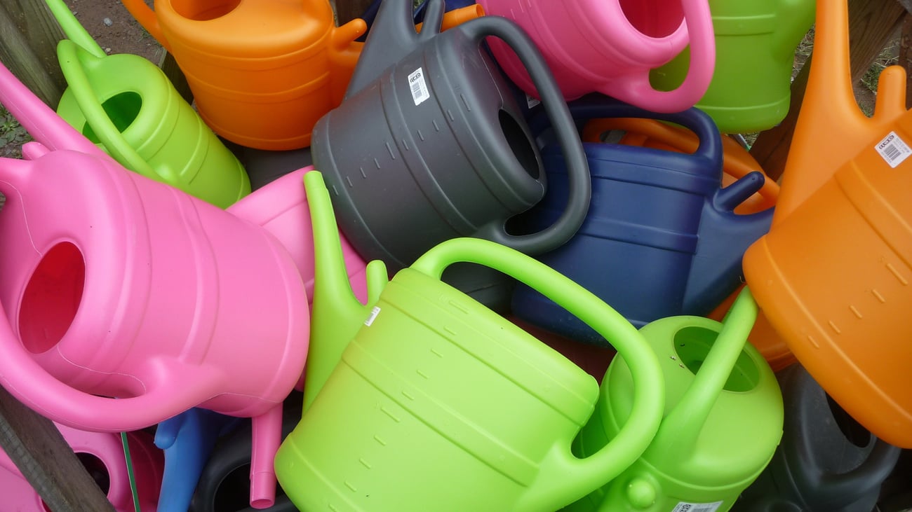 durcheinander liegende große Gießkannen in den Farben hellgrün, orange, dunkelgrau, blau und rosa