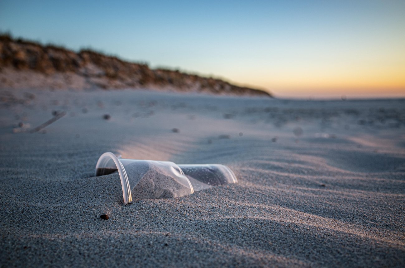 Blick auf einen halb im Sand vergrabenen farblosen Plastikbecher, im Hintergrund unscharf sandbewachsene niedrige Dünen, Sonnenuntergangsstimmung am Meer.