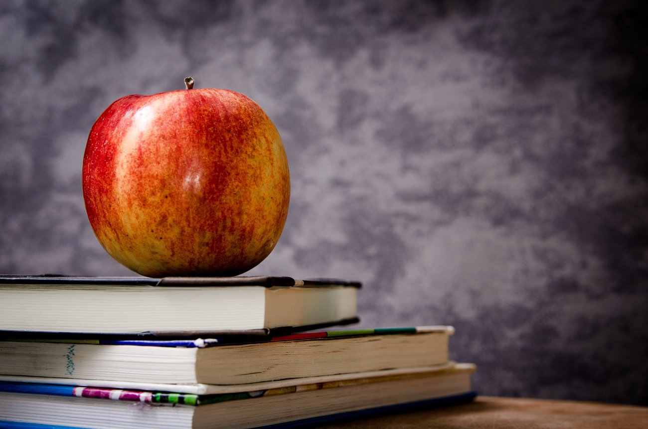 Ein rot-gelber Apfel liegt auf mehreren Büchern, die teilweise schräg übereinander gelegt wurden. Die Bücher liegen auf einer Holzplatte. Im Hintergrund ist eine hellgrau-dunkelgrau marmorierte Wand.
