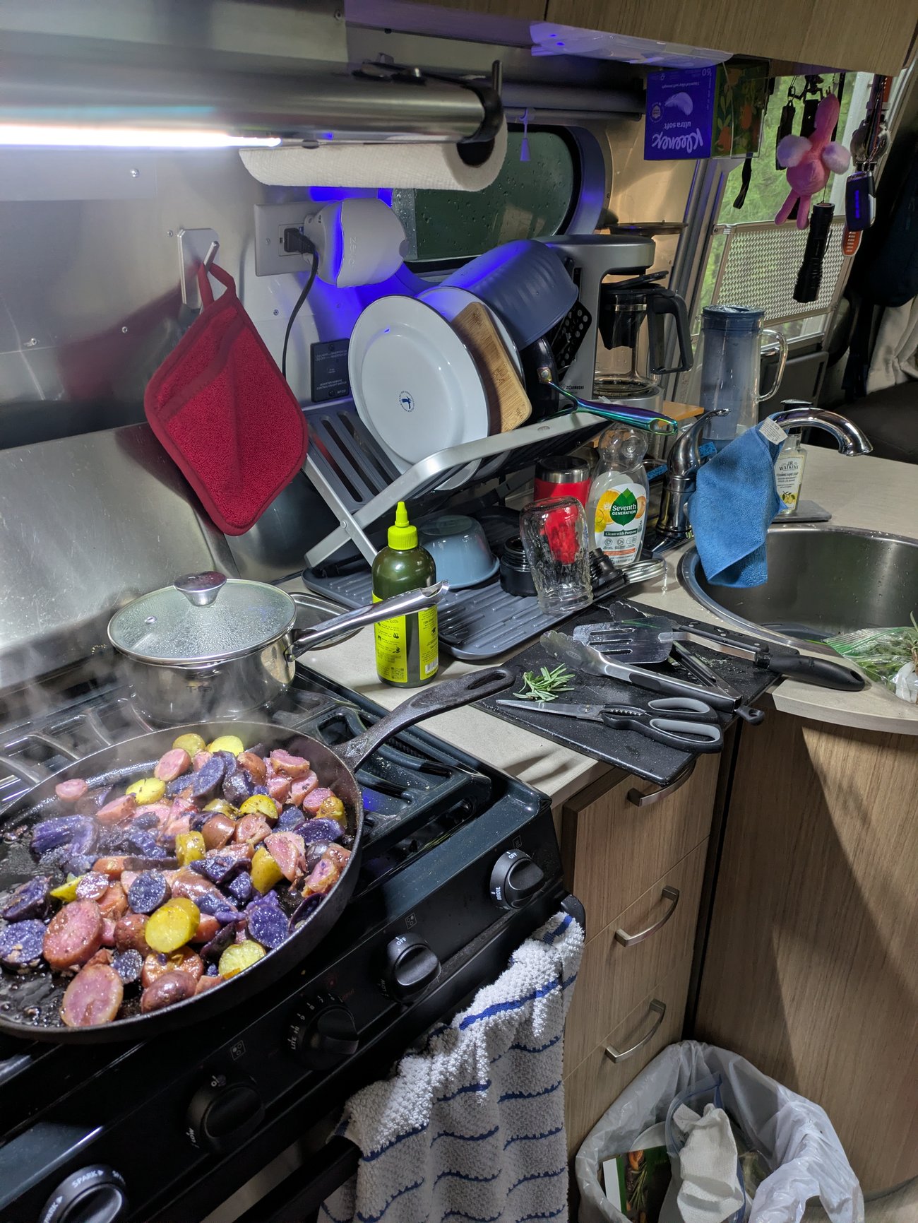 Compact Airstream kitchen with a meal cooking on the stove, dishes drying by the sink, and limited counter space in use.