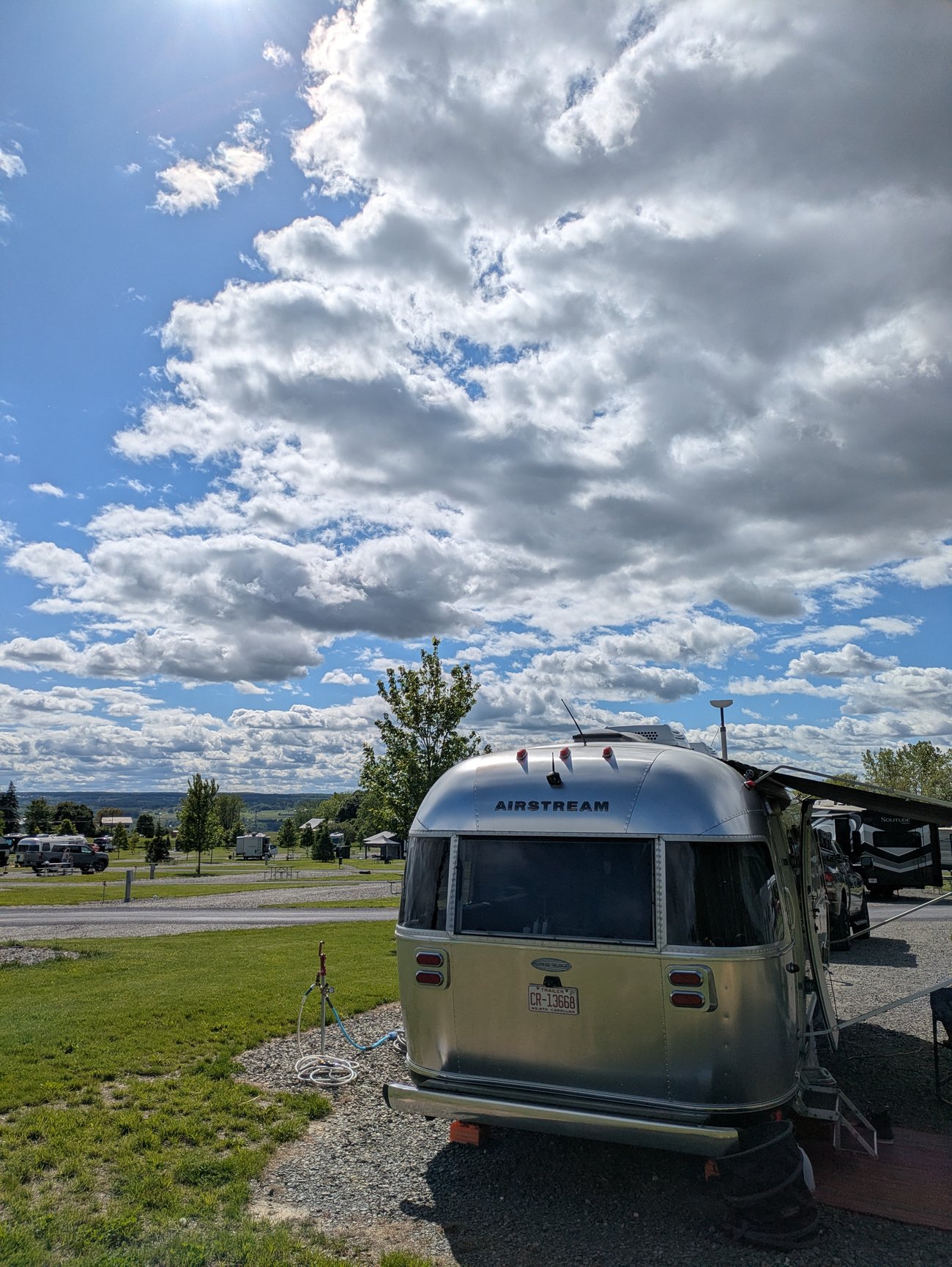 Airstream trailer parked at a campground beneath a wide sky filled with dramatic summer clouds, with rolling hills visible in the distance.