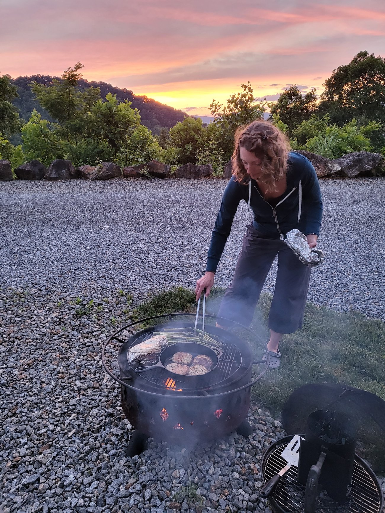 Abby cooking over a campground fire pit at sunset, with rolling hills and a colorful evening sky in the background.