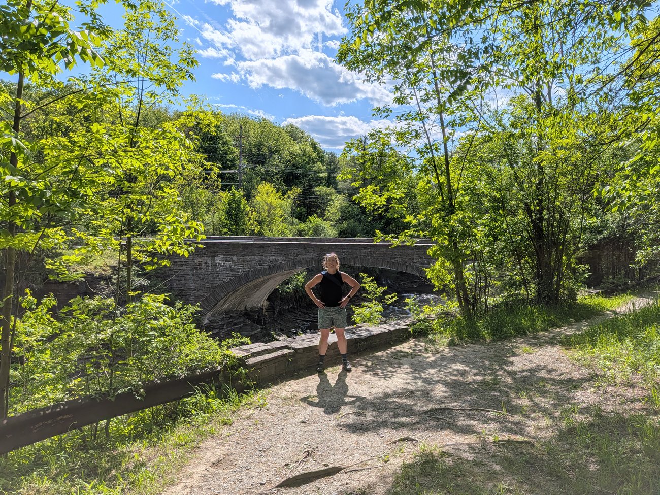 Abby standing on a wooded trail in front of a historic stone arch bridge on a sunny day.