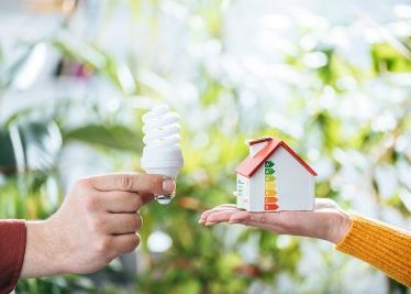 Two hands holding a small model house and a light bulb against a blurred green background