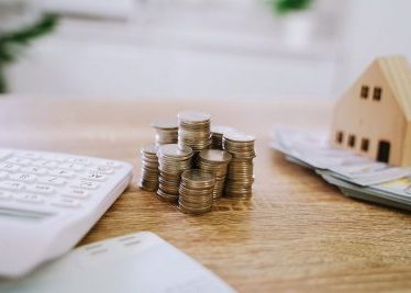 Stacks of coins on a wooden desk beside a calculator, papers, and a small house model