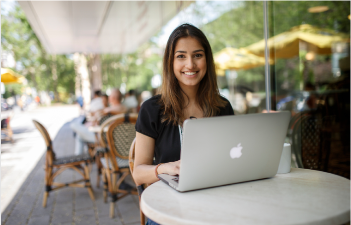 Female student, smiling. Seated outside at table with computer. 