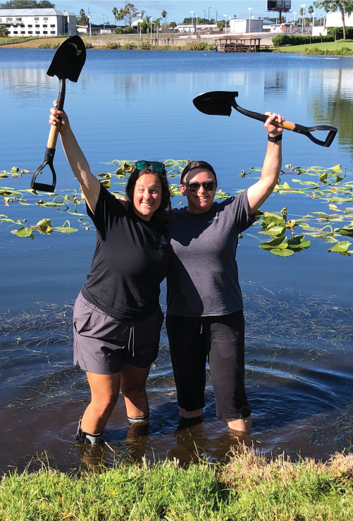 2 women wading in lake holding shovels above their heads