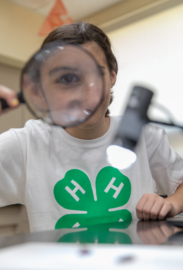 youth wearing 4-H logo shirt sitting at table holding magnifying glass up to eye